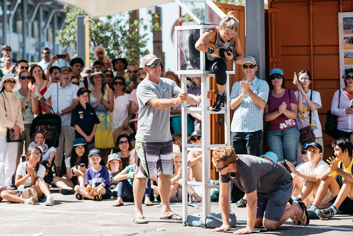 Street performer folding herself into a small box, with a crowd of people watching. 